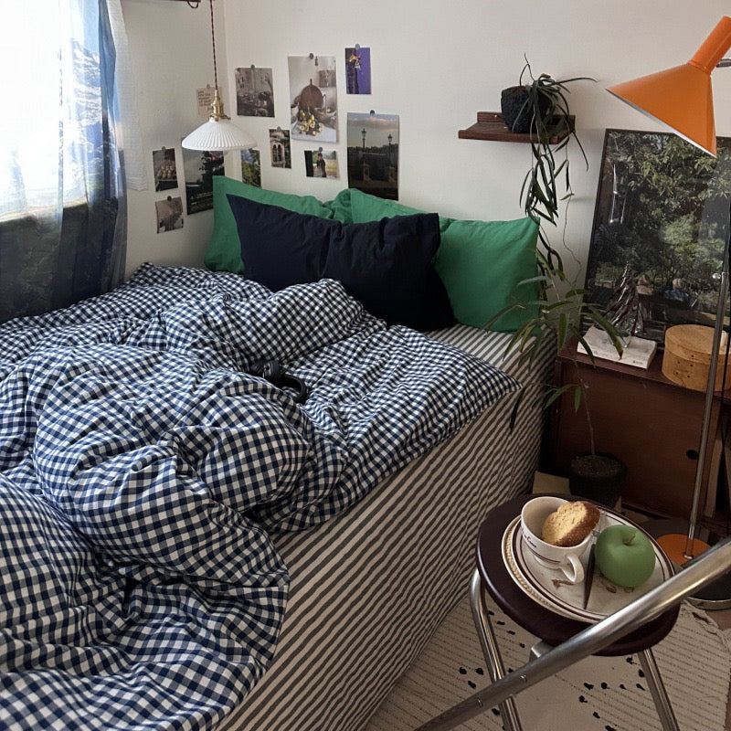 A cozy bedroom scene with a bed made up in blue-and-white gingham bedding, striped sheets, and green pillows, in a real room with a side table, lamp, plant, and wall decor.