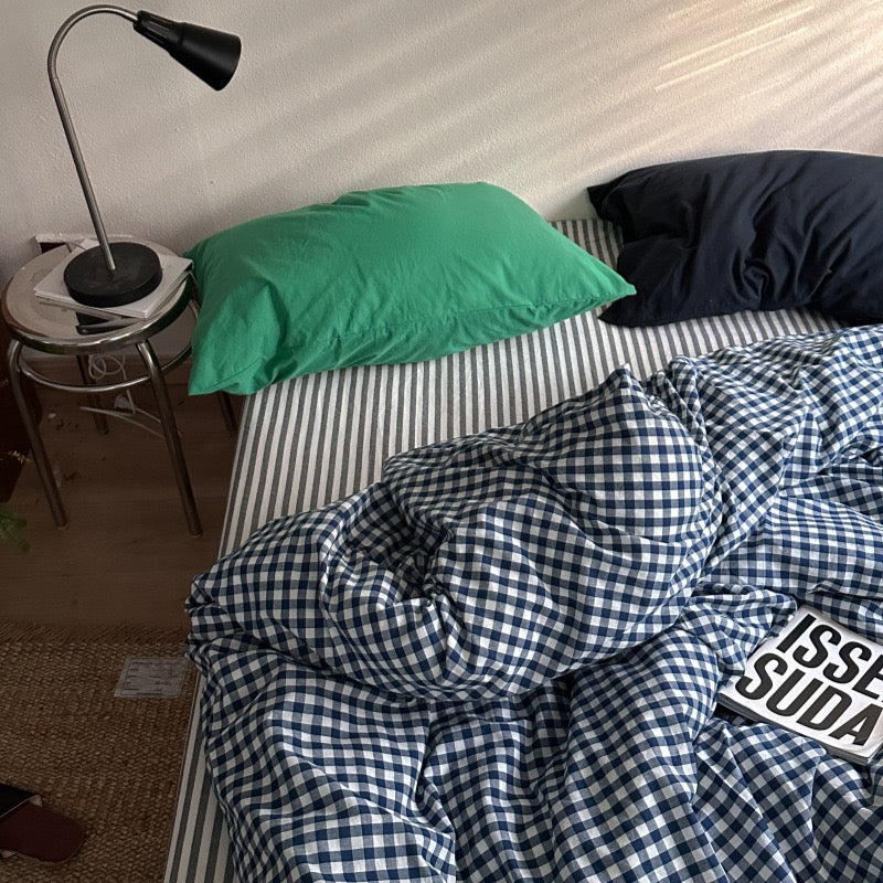A bed with a blue and white gingham duvet cover and striped sheets, featuring a green pillow and a dark navy pillow, with a black metal desk lamp on a side table next to it.