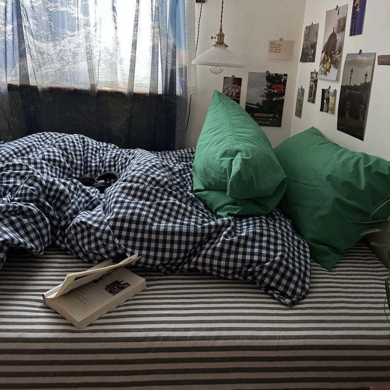 A bed in a real bedroom setting with rumpled blue-and-white gingham bedding, two green pillows, a striped sheet, a hanging lamp, wall photos, and a book on the bed.