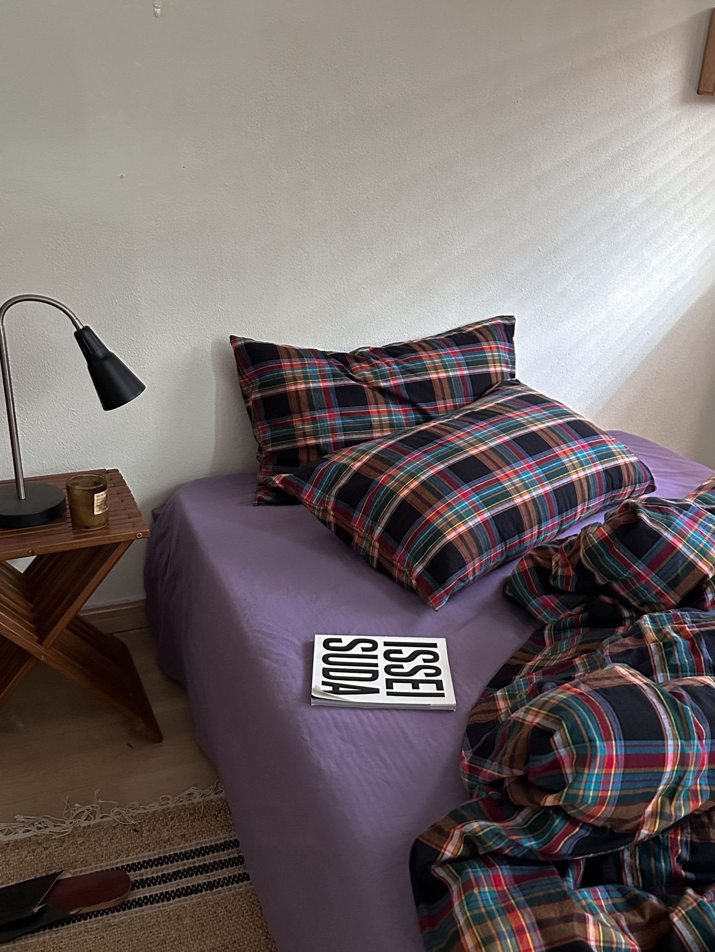 A bed styled in a bedroom setting with a purple sheet and matching plaid bedding in black, red, green, and blue. A wooden side table with a black lamp sits next to the bed.