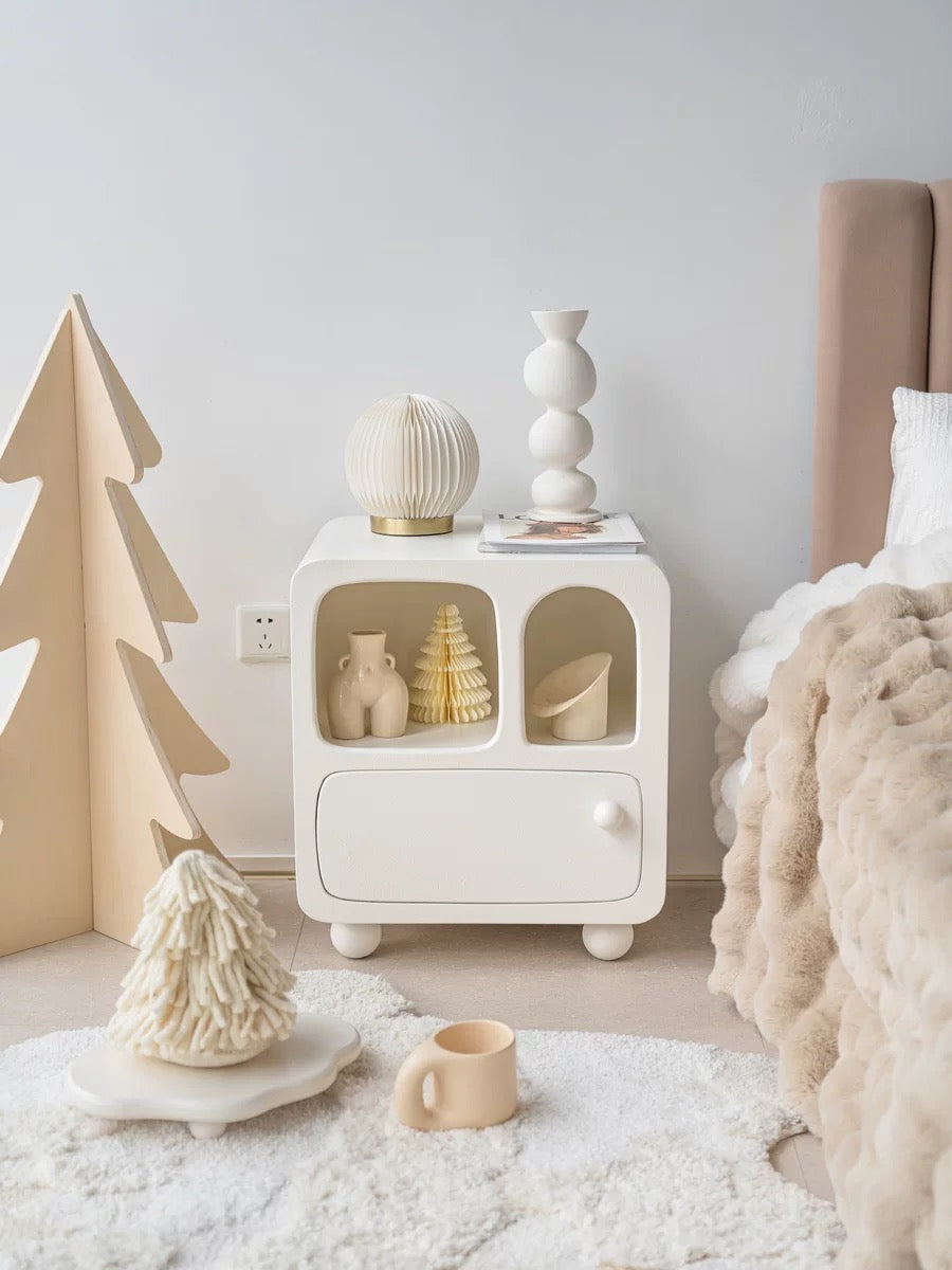 A white nightstand with shelves and a lower drawer stands beside a bed, with small vases in the openings.