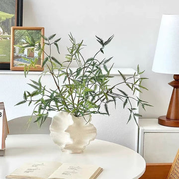 Decorative plant in a white vase on a table with a lamp and books in the background.
