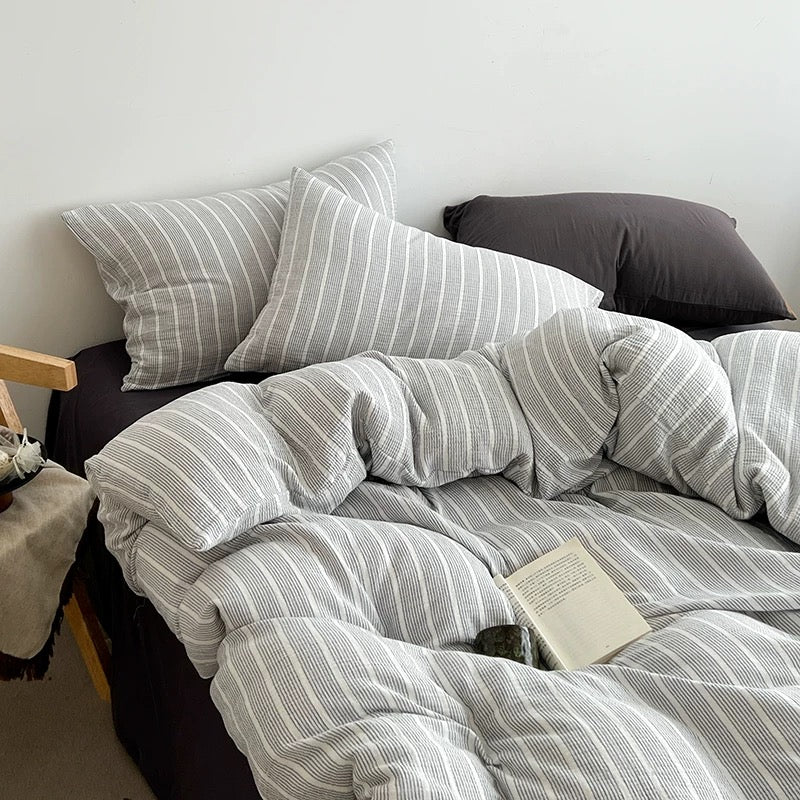 A cozy bed setup with a light gray and white striped bedding set, including a duvet cover and two pillows, styled in a bedroom setting with a dark headboard and a book on the bedding.