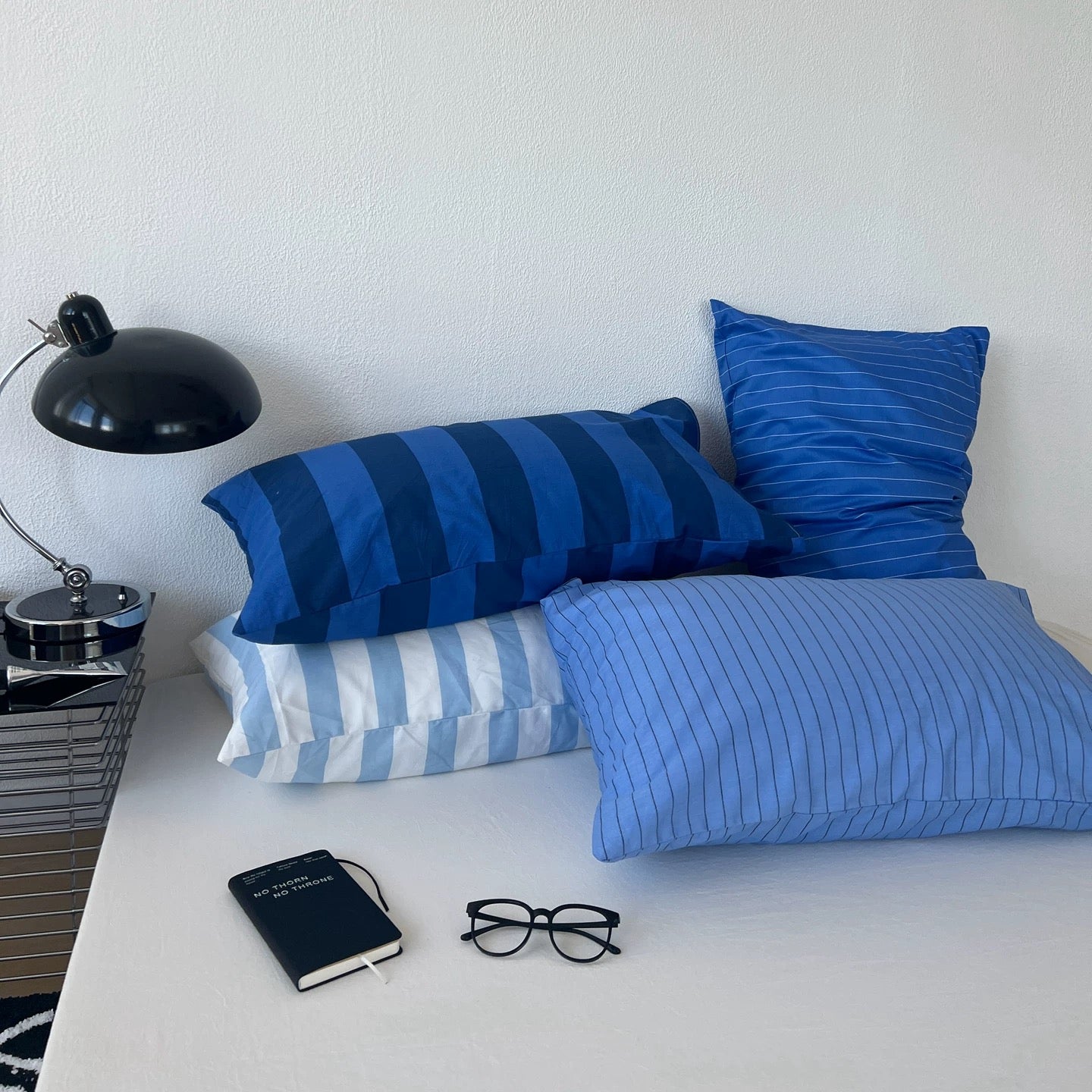 Three blue striped and solid blue pillows arranged on a white bed alongside a black desk lamp, notebook, and eyeglasses against a white wall.