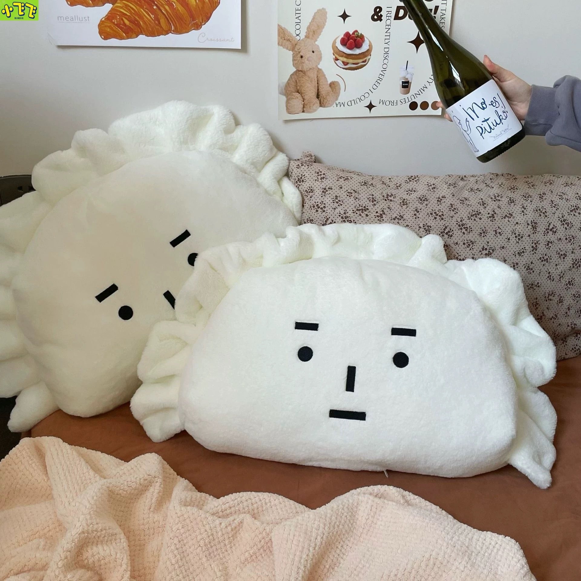 Two white plush toys with face designs on a bed, with a bottle of wine in the background.