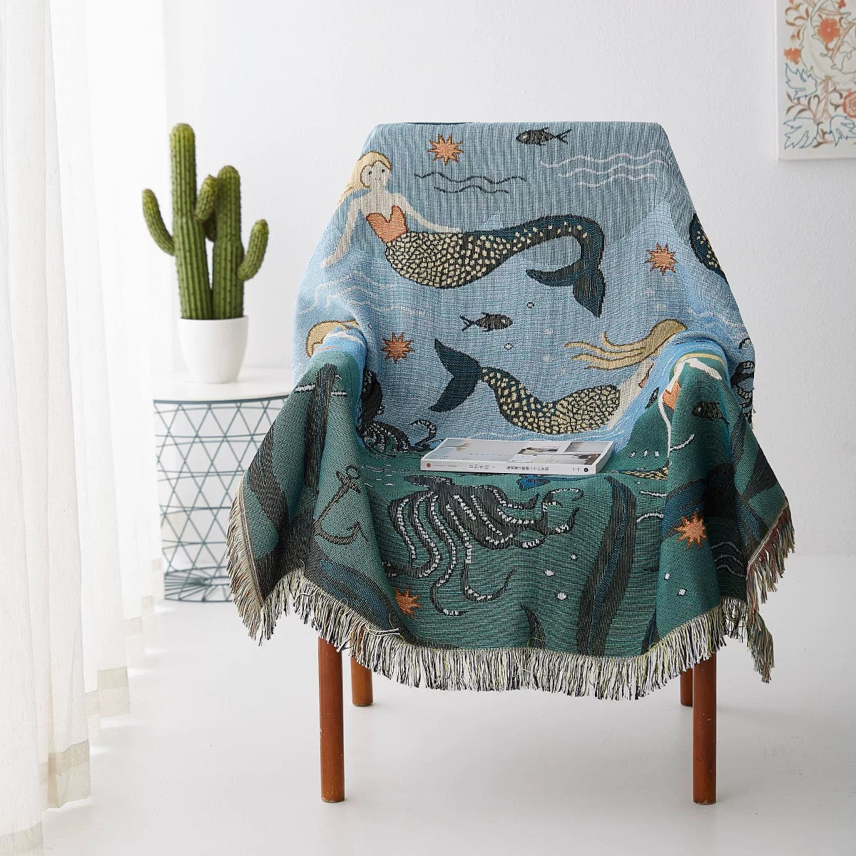 Decorative blanket with mermaid and sea creature design draped over a chair in a room with a cactus plant.
