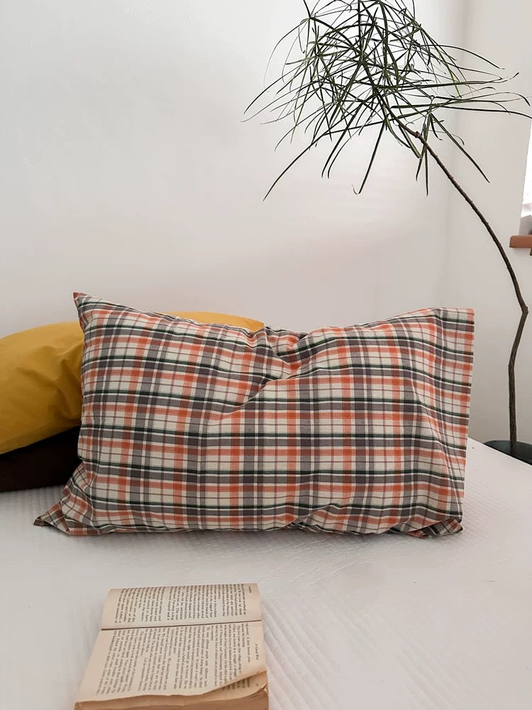 An orange plaid pillowcase lies on the bed beside a plant and an open book.