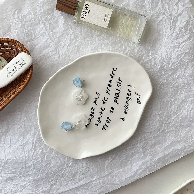 An organic-shaped white ceramic dish with handwritten black French text, styled on a crumpled white paper background with a perfume bottle, a small basket, and a white candle nearby.