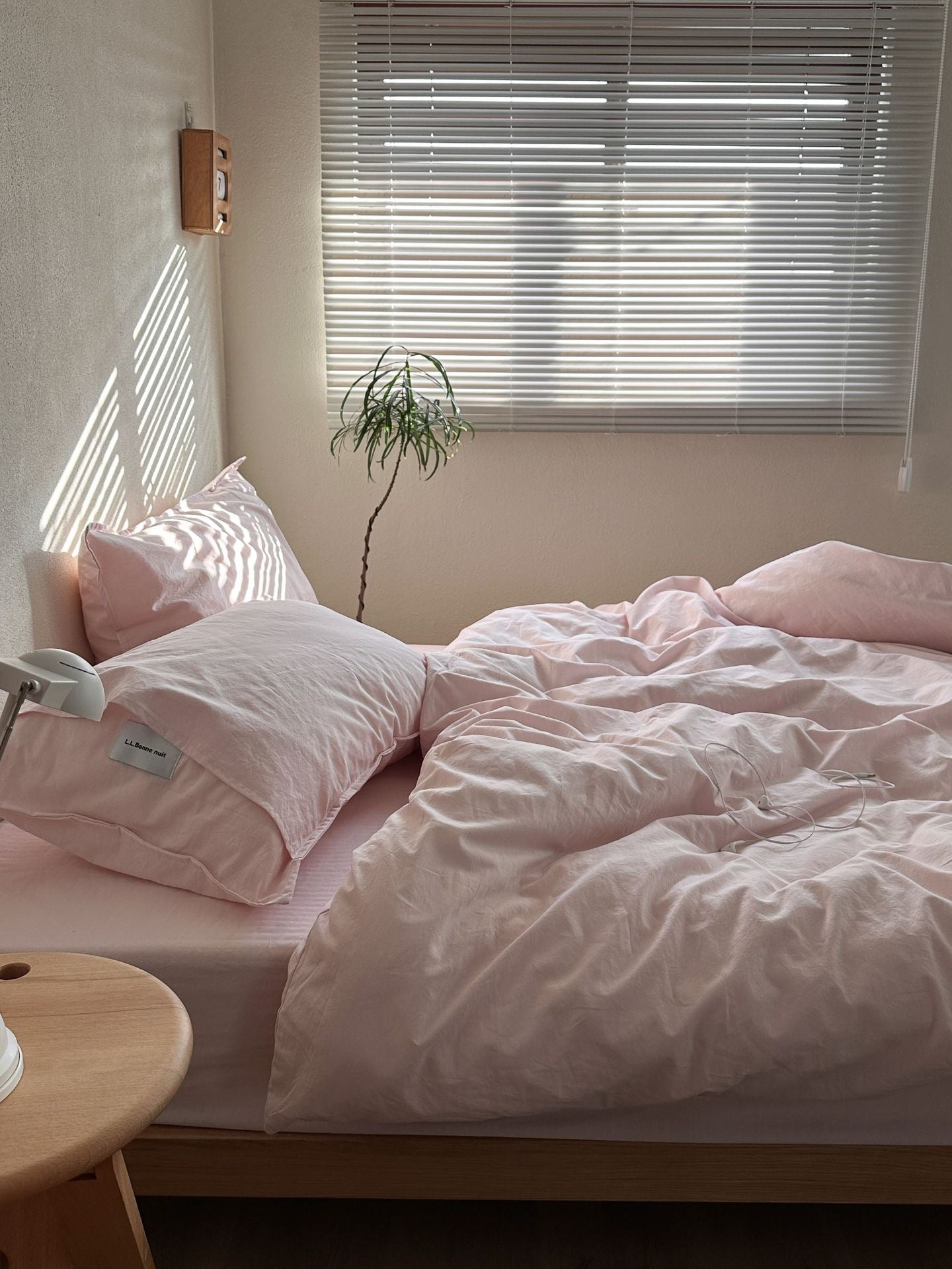 A neatly made bed with light pink bedding and matching pillows in a softly lit bedroom with white window blinds and a small plant.
