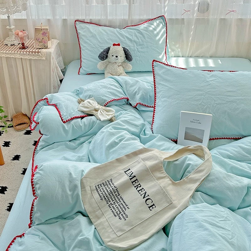 A light blue bedding set with red trim on a bed, styled with a plush dog pillow, a cream canvas tote bag, and a small card, in a bright bedroom setting.