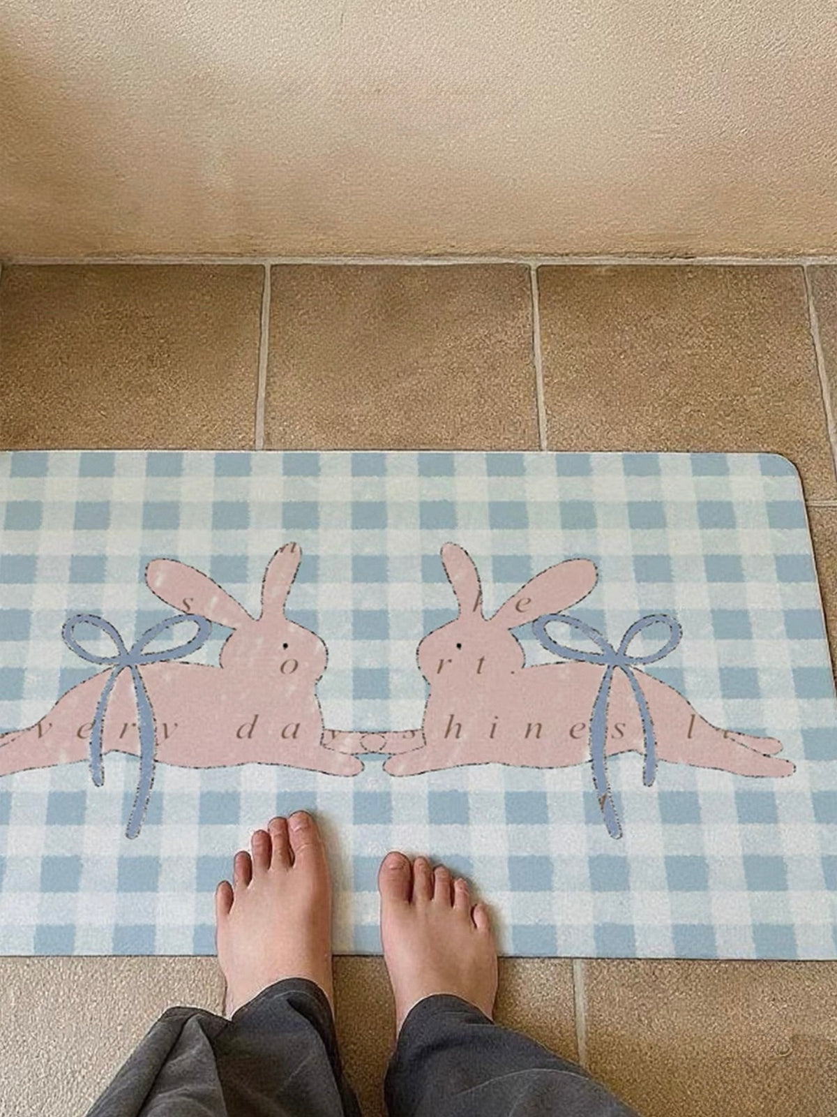 A pastel doormat beneath bare feet shows pink bunnies, blue bows and a blue and white gingham background.
