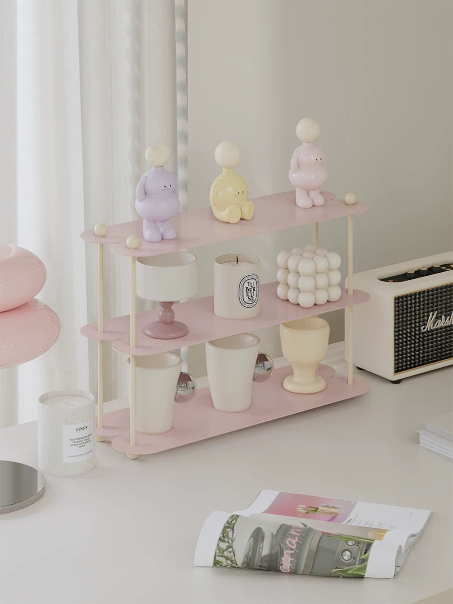 A pink pastel shelf with cream posts and round ball details displays mugs, candles and small figures.
