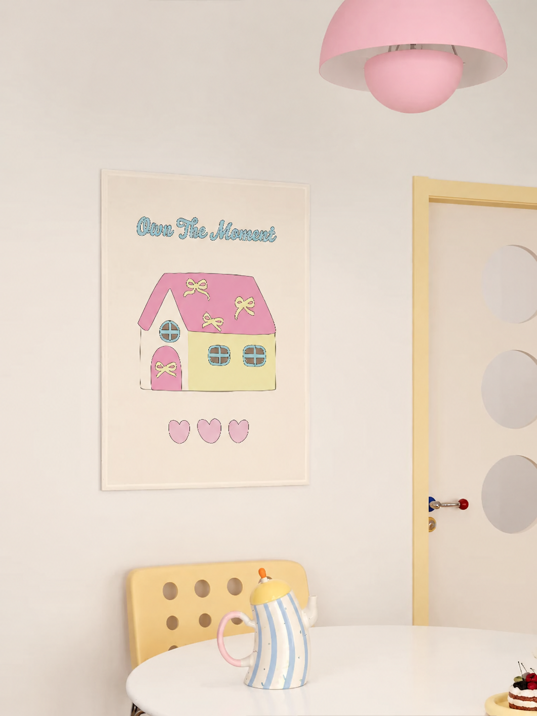 A pastel tapestry with a pink roof and pale yellow house hangs above a striped teapot and yellow chair.
