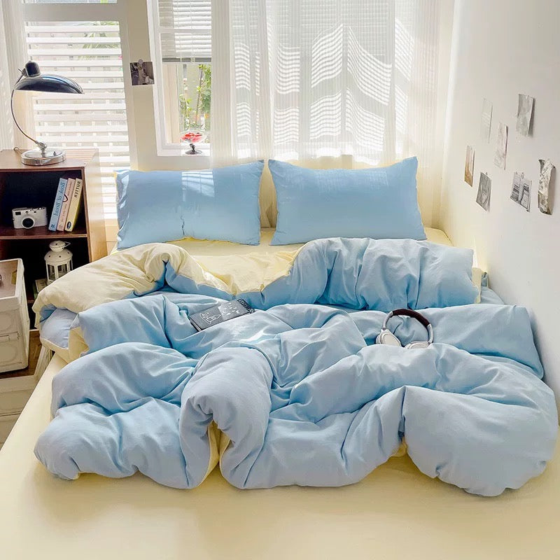Light blue bedding set on a bed in a bright room with windows and shelves.