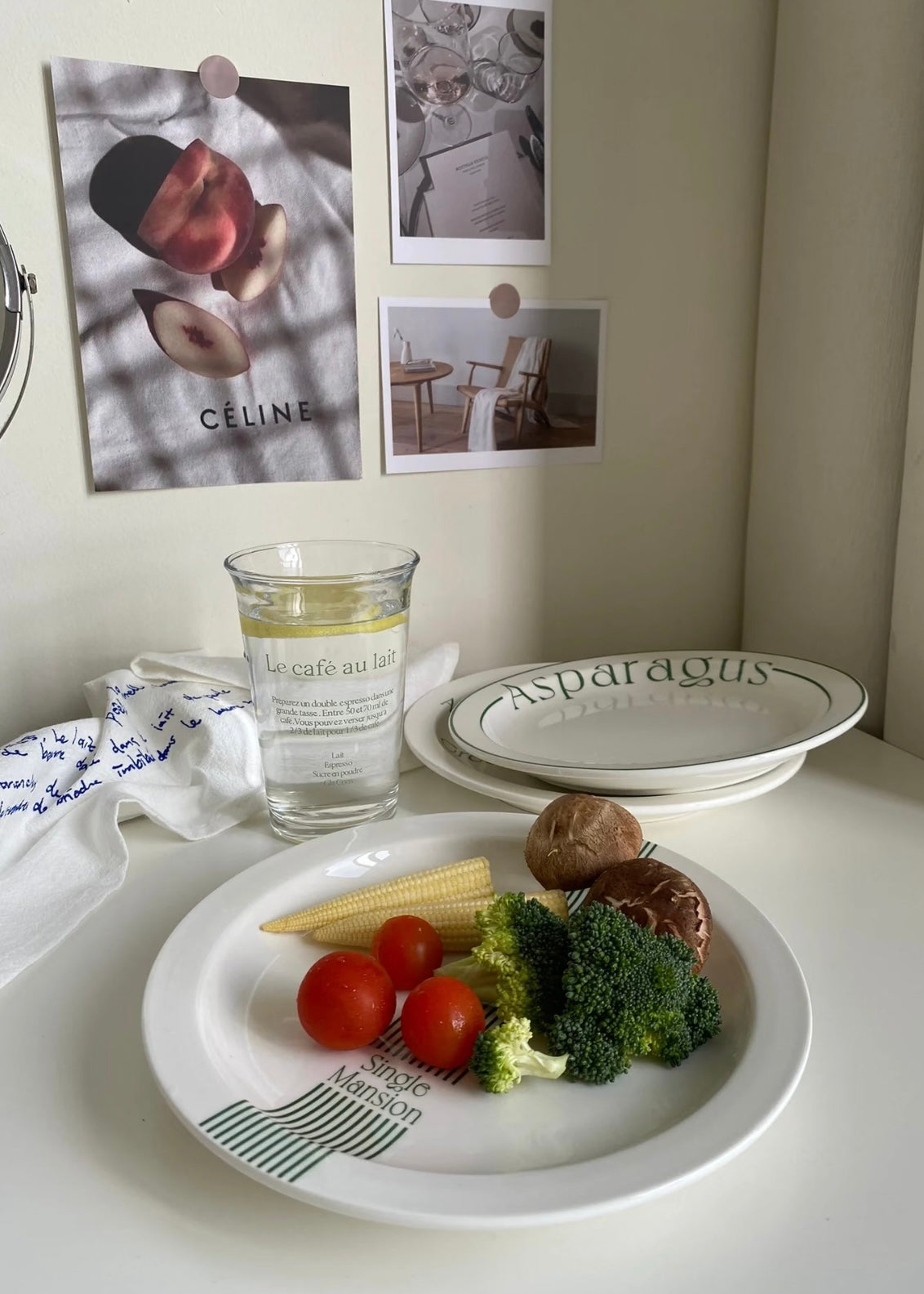 A set of white ceramic plates with green printed text and stripes, shown on a tabletop with food props and a candle. One plate is labeled 'Asparagus' and another has 'Maison' printed near the rim.