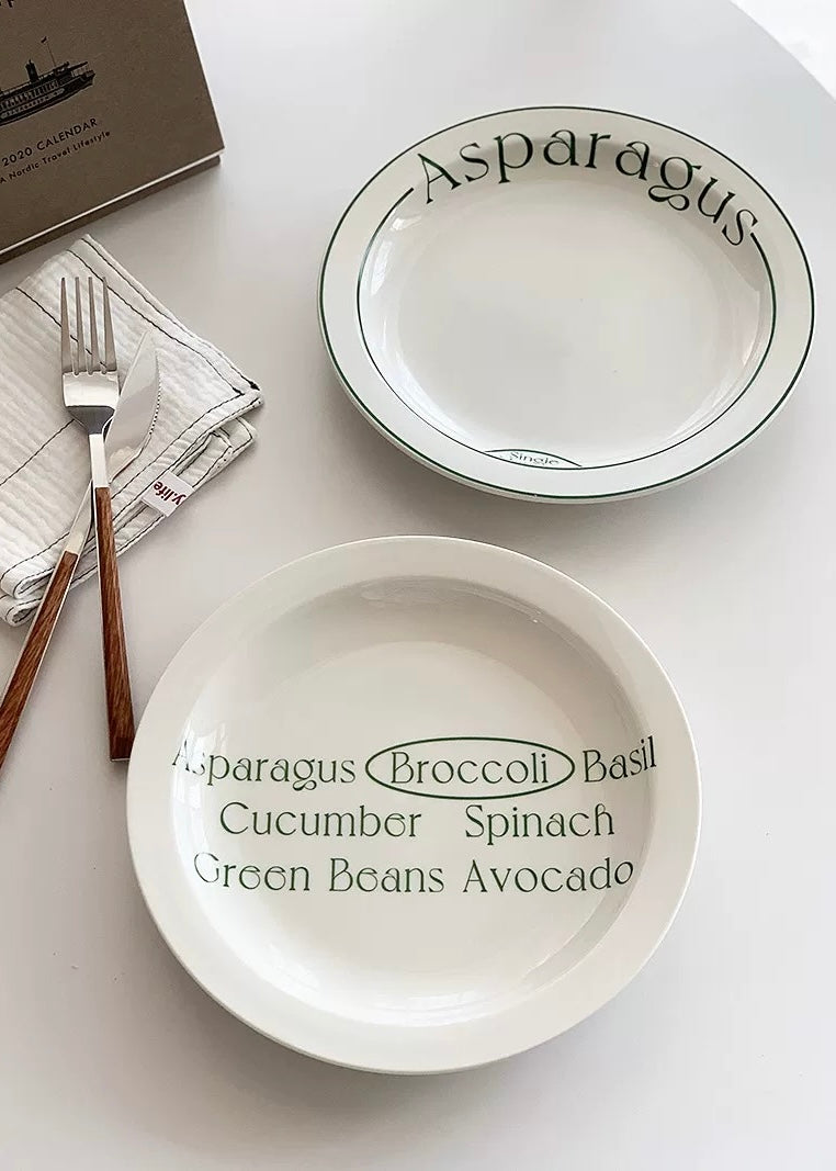 Two white ceramic plates with green printed text and borders, shown on a light tabletop with a fork, folded napkin, and a book corner visible.