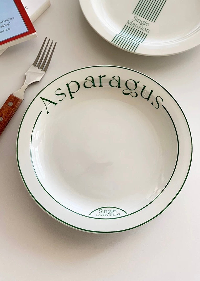A white ceramic serving bowl with 'Asparagus' printed in dark green script and border lines, shown on a light surface with a fork and part of another plate visible.