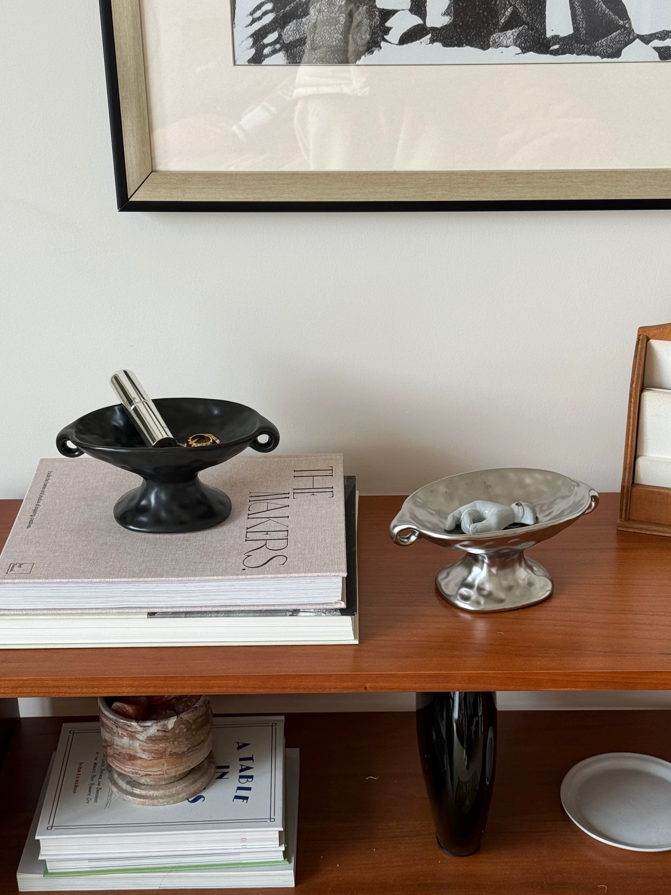 A styled interior scene showing a wooden shelf with decorative objects, including a black bowl-shaped dish on stacked books, a silver pedestal bowl, and framed wall art above.