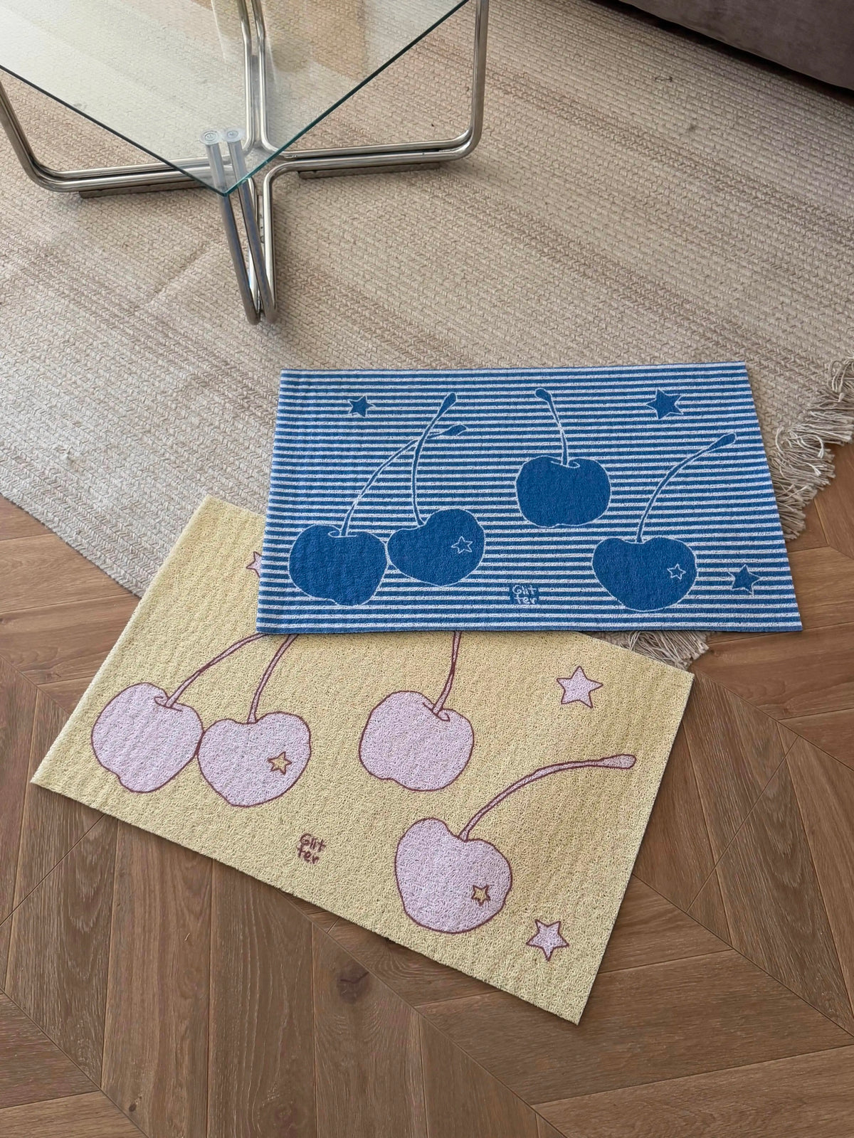 Two rectangular floor mats with cherry designs, one blue and white striped with dark blue cherries, and one beige with light pink cherries, displayed on a wooden parquet floor.