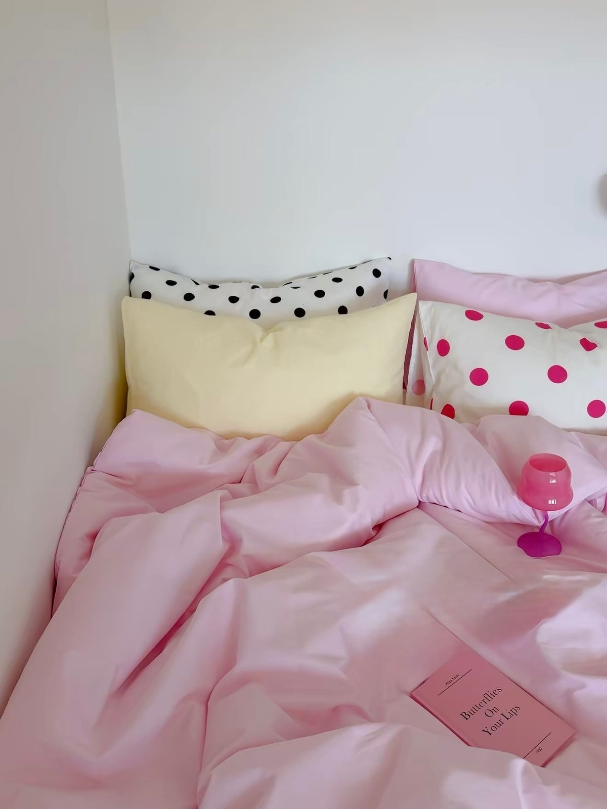 Close view of a pink bed with a pale yellow pillow, black and fuchsia dot pillowcases, and a metal bedside lamp.