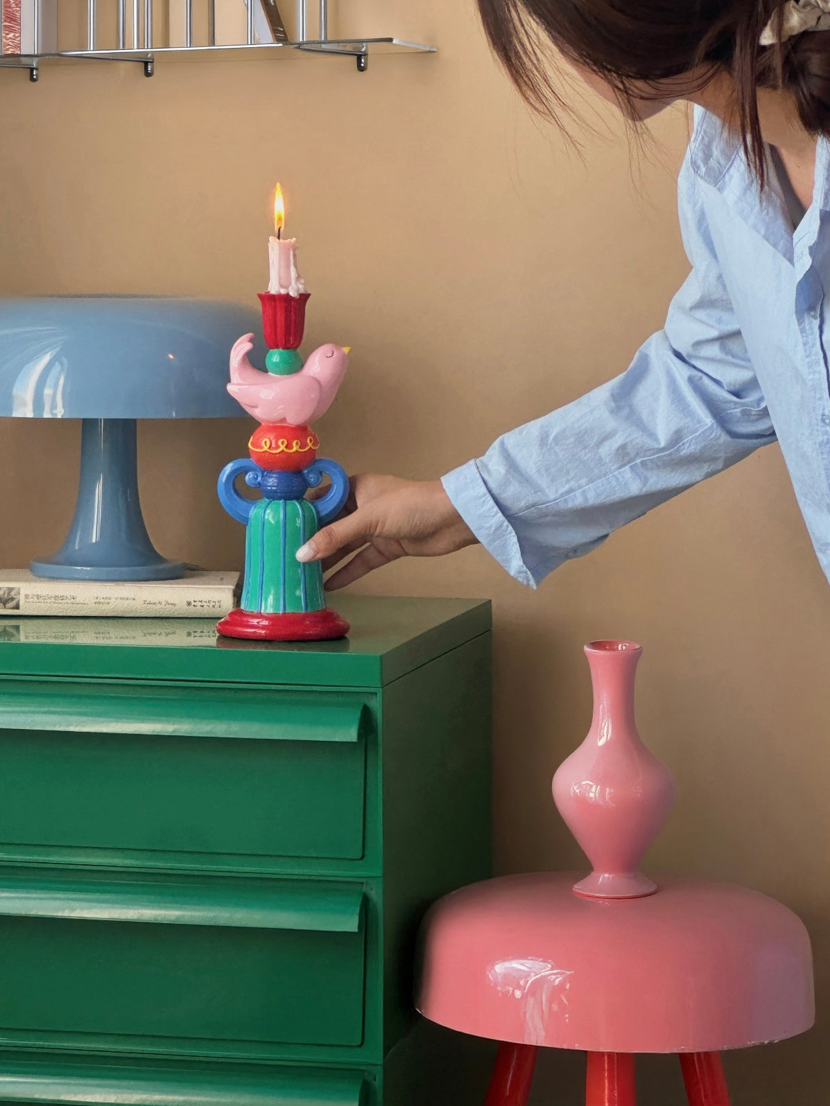 Pink bird candle holder with a lit candle placed on a green cabinet beside a blue table lamp.