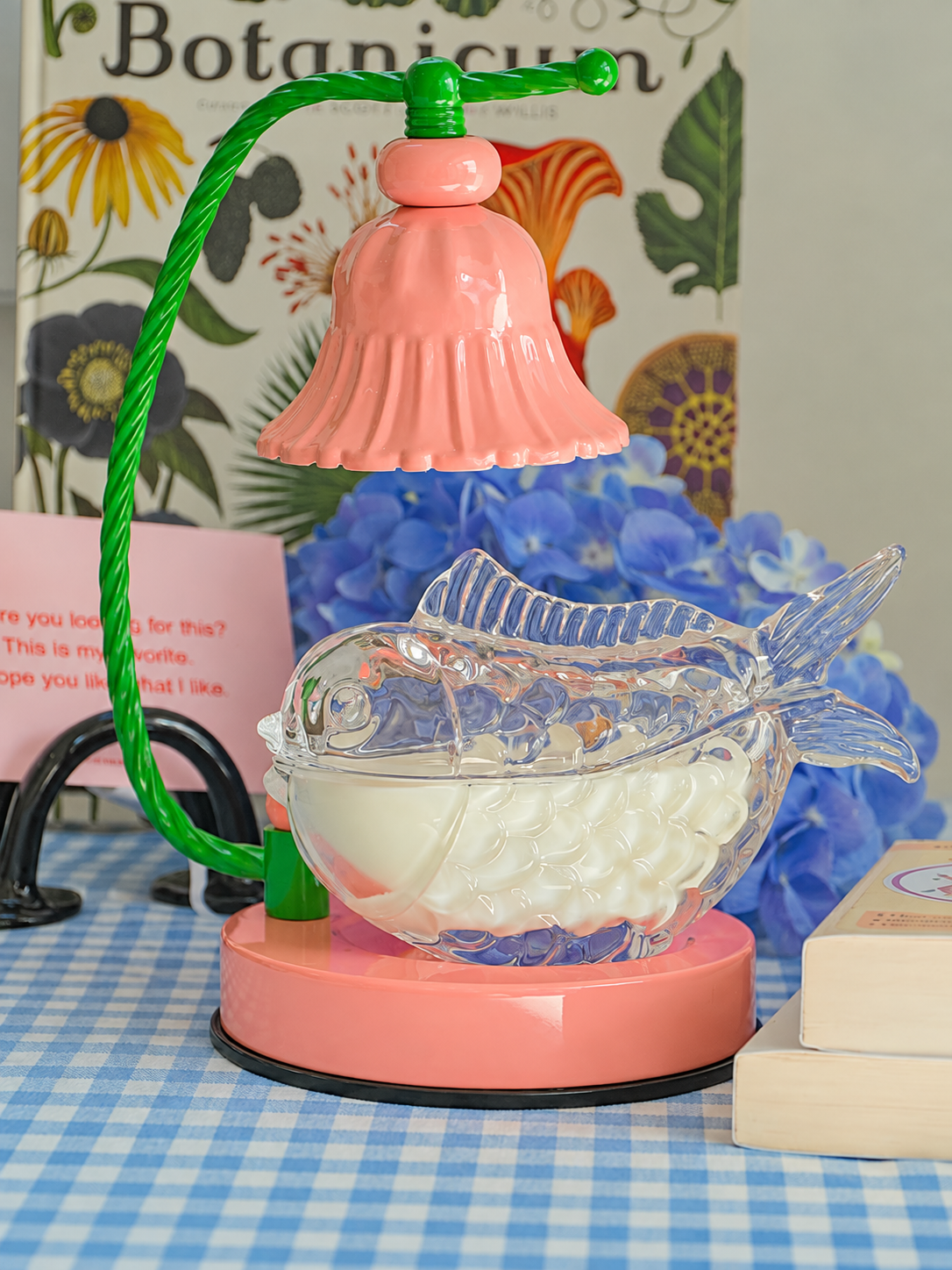 A decorative fish-shaped glass bowl filled with white wax melts, set on a pink circular base with a green wire arm and pink bell-shaped lamp shade, styled on a blue-and-white checkered tablecloth with flowers and a botanical book in the background.