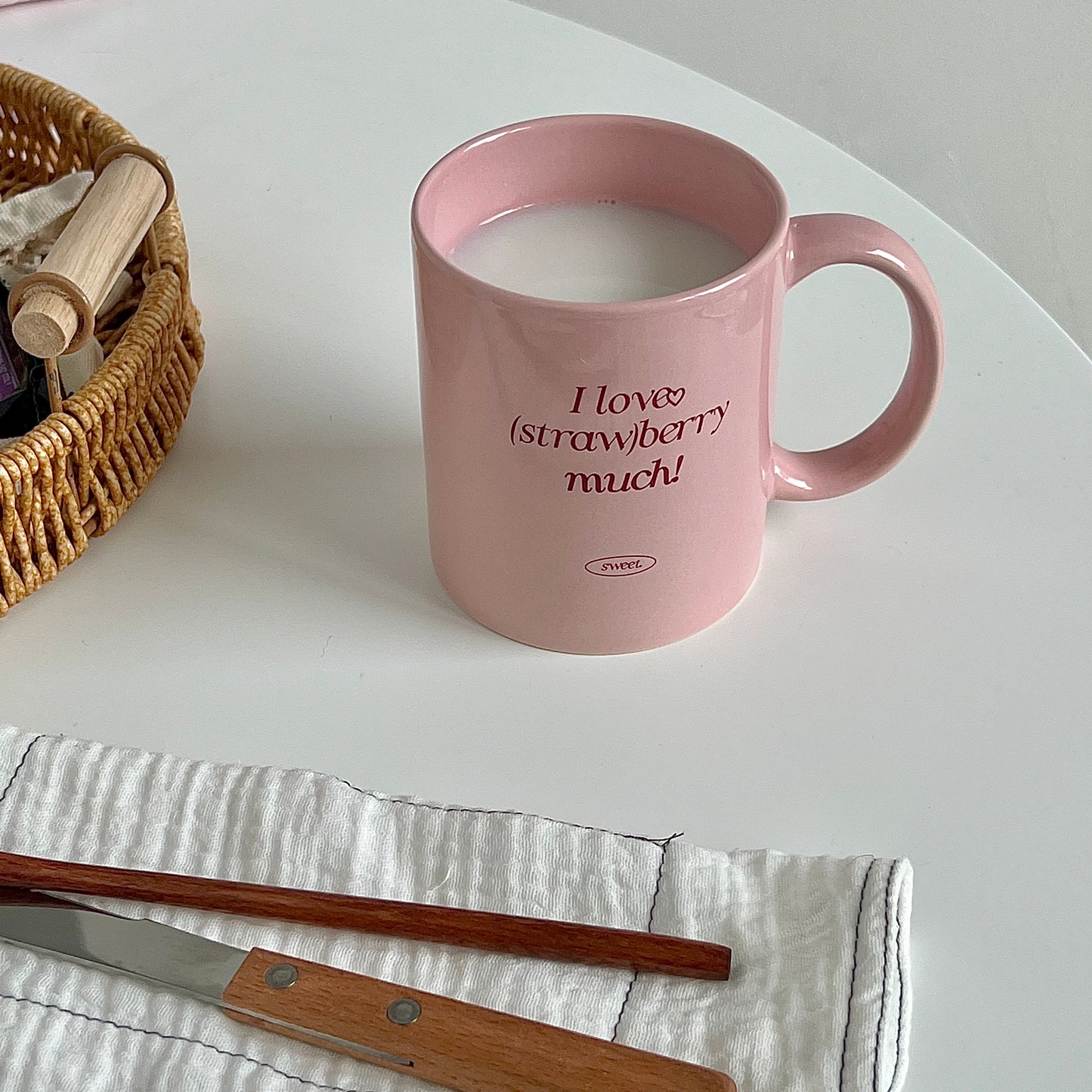 A pink ceramic mug filled with a light pink drink sits on a white tabletop. The mug has the phrase 'I love (strawberry much!)' printed in dark pink text with a small strawberry icon.