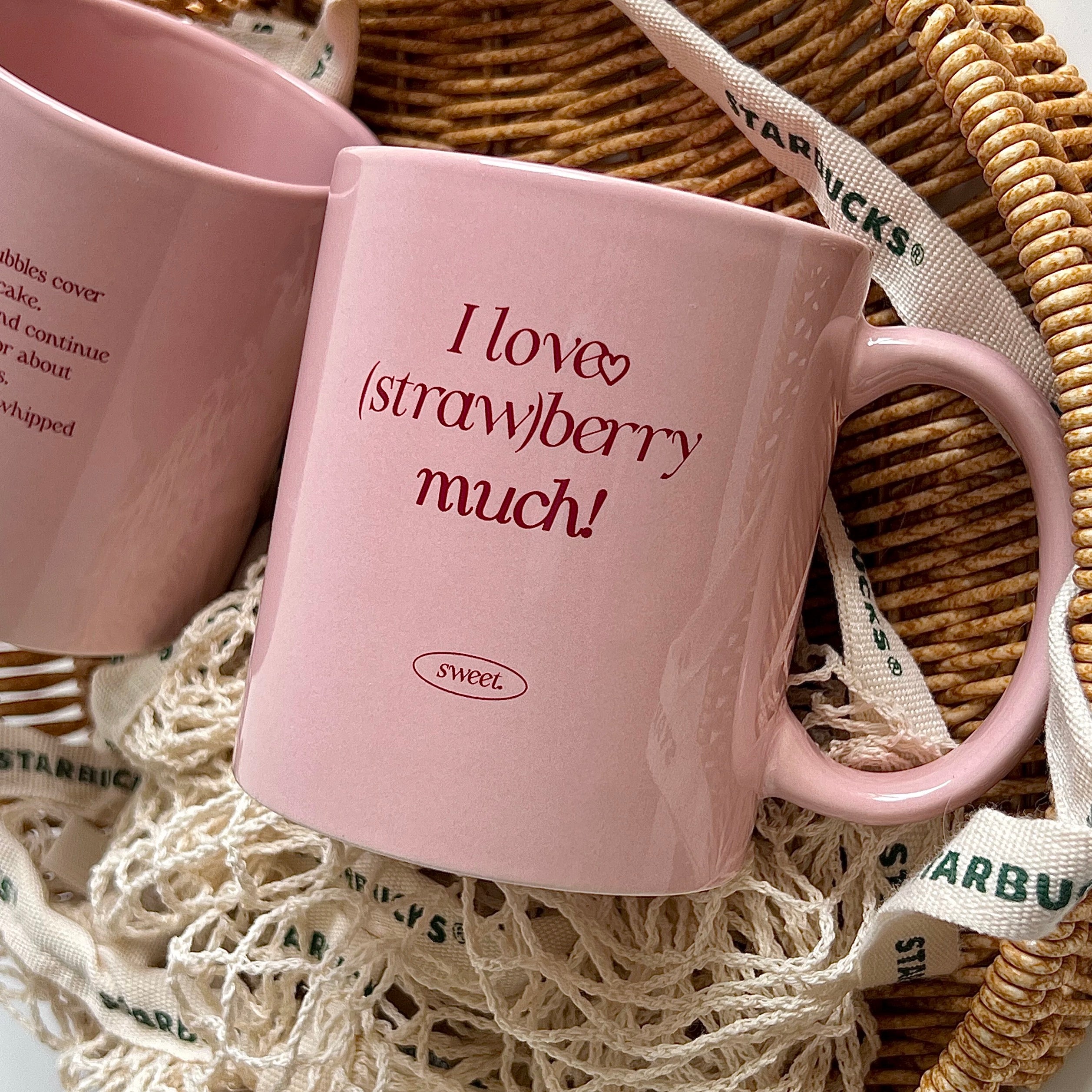 A pink ceramic mug with the text 'I love (straw)berry much!' in dark red lettering, displayed inside a woven wicker basket with cream-colored netting and a printed ribbon. Another pink mug is partially visible in the background.