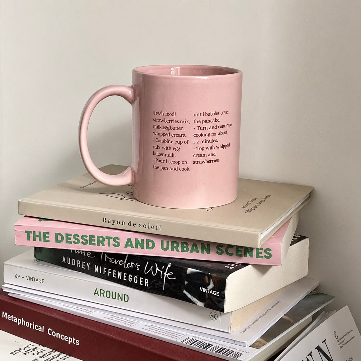 A light pink ceramic mug with black printed text sits on top of a stack of books against a plain off-white wall.