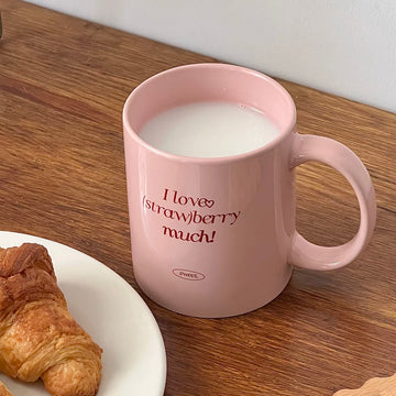 A pink ceramic mug filled with milk sits on a wooden table next to a plate with a croissant. The mug has the text 'I love strawberry much!' printed in red, with a small oval logo below that reads 'SWEET'.