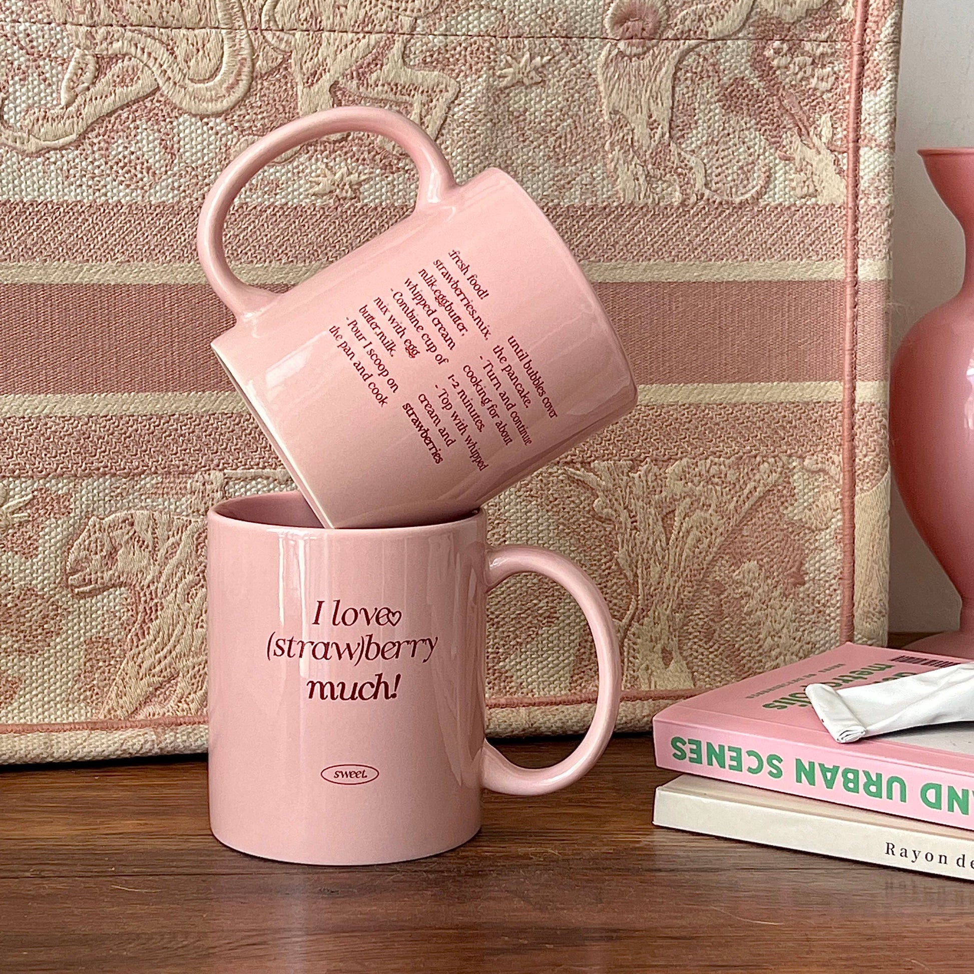 Two pink ceramic mugs on a wooden table in front of a patterned fabric backdrop. One mug is stacked on top of the other, with the top mug tilted to show printed text.