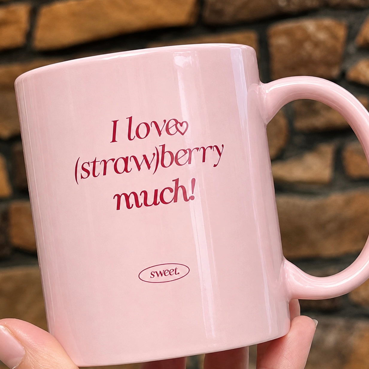 A light pink ceramic coffee mug with the text 'I love (straw)berry much!' and a small 'sweet.' logo printed in dark pink, held by a hand against a blurred brick wall background.