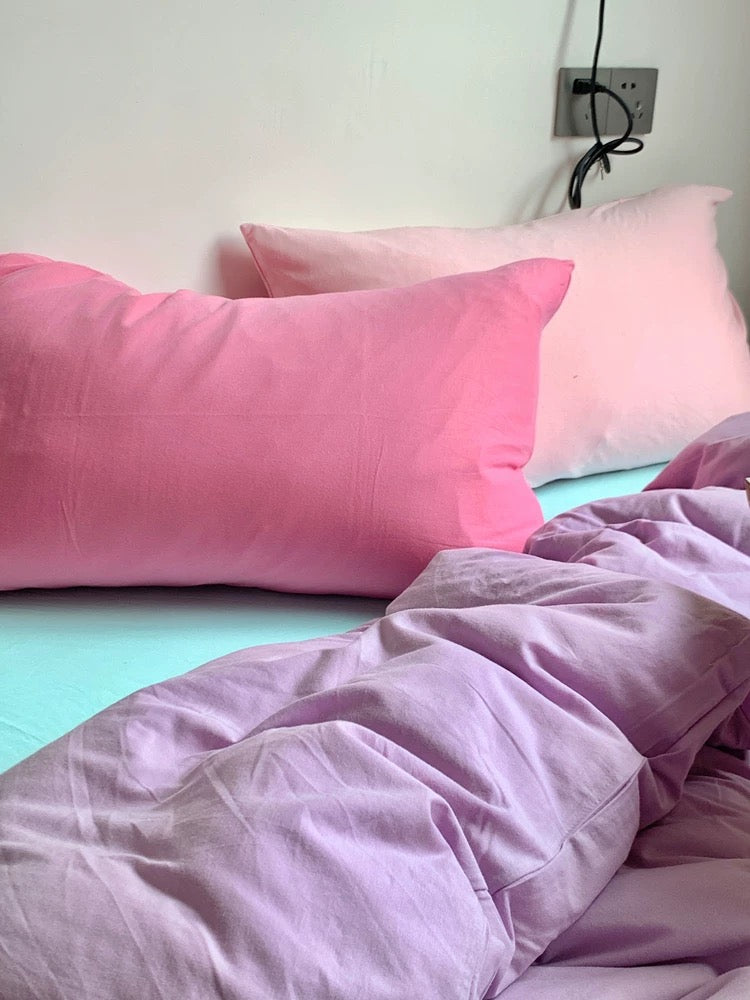 Pink pillows and purple comforter on a bed with a light blue wall in the background.