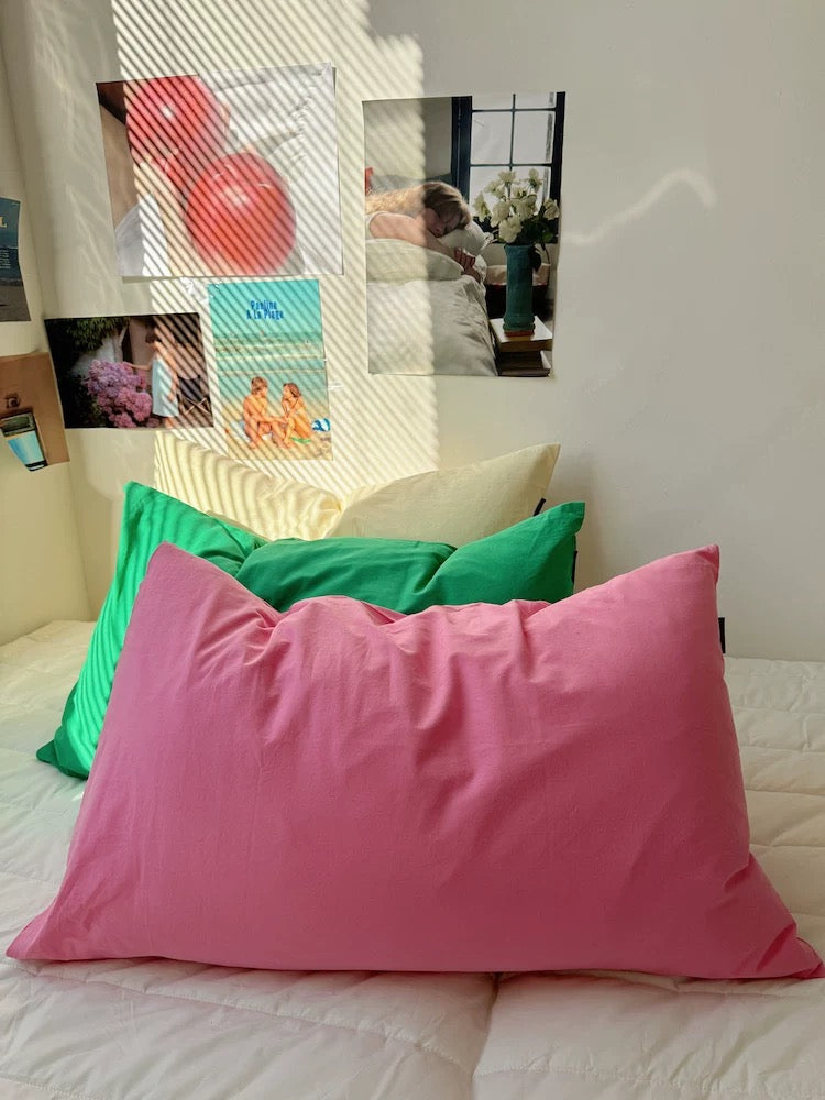 A pink pillowcase on a bed in a bedroom setting, with green and cream pillows behind it and framed wall art in the background.