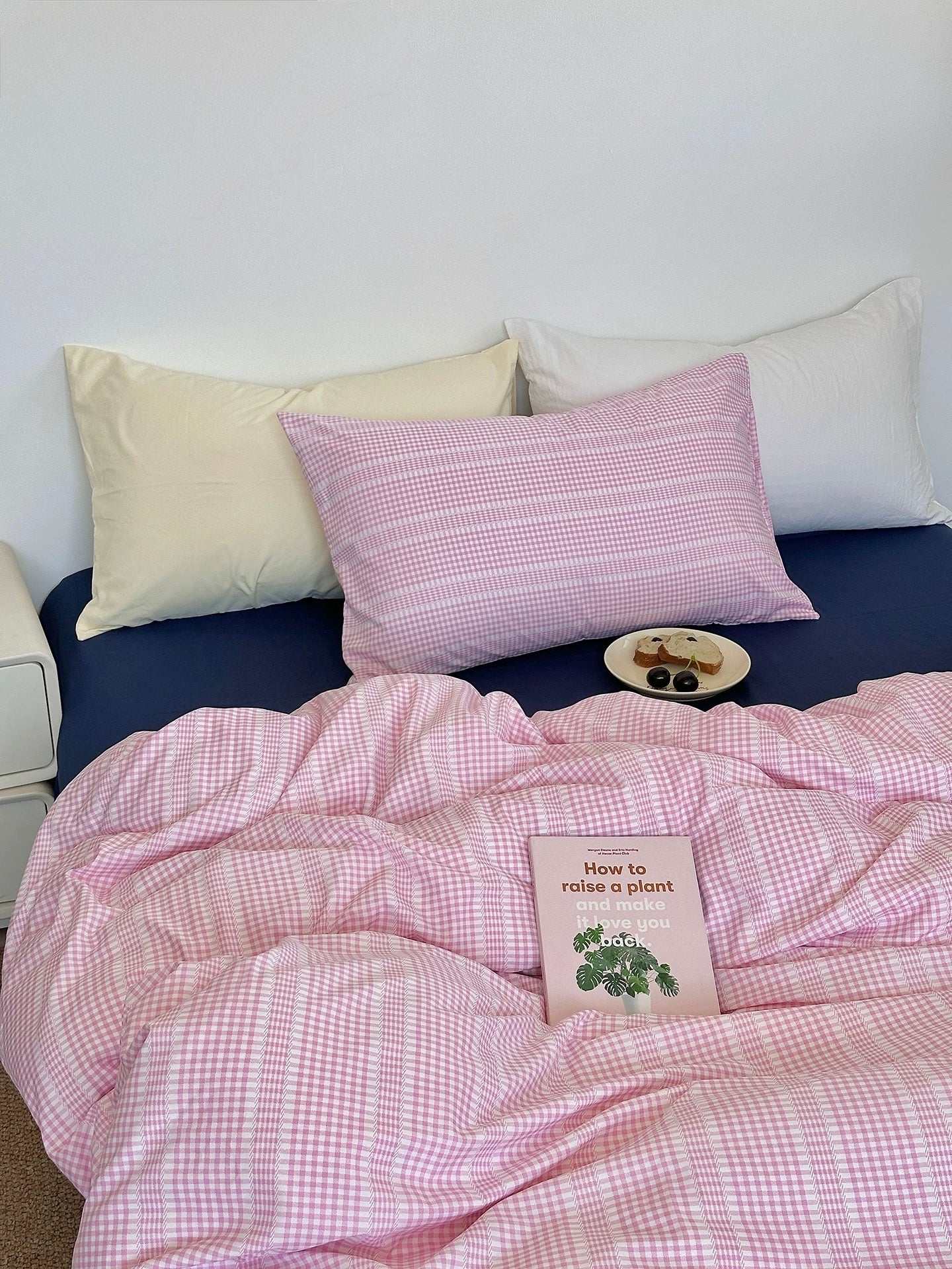 Pink checkered comforter on a bed with pillows and a book titled 'How to grow a plant'.