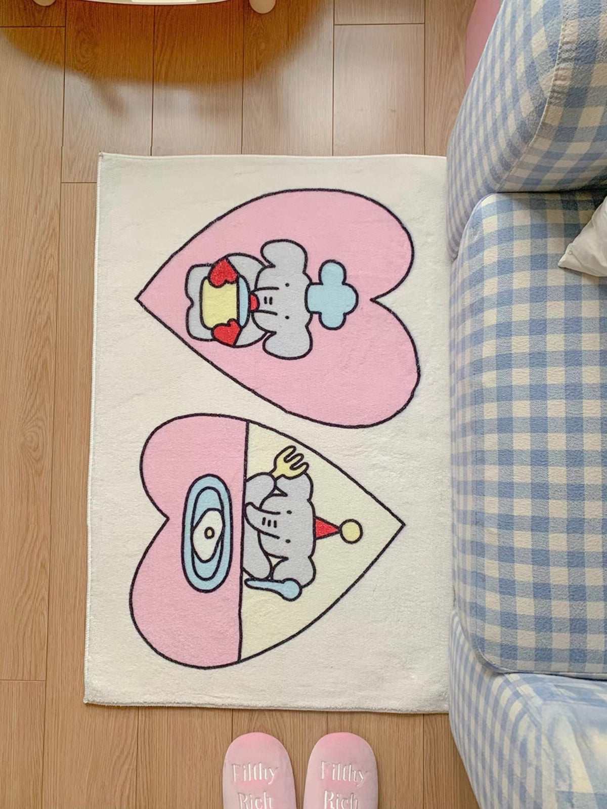 A pink heart rug lies beside a checked chair, with one elephant cooking and another seated above a plate.