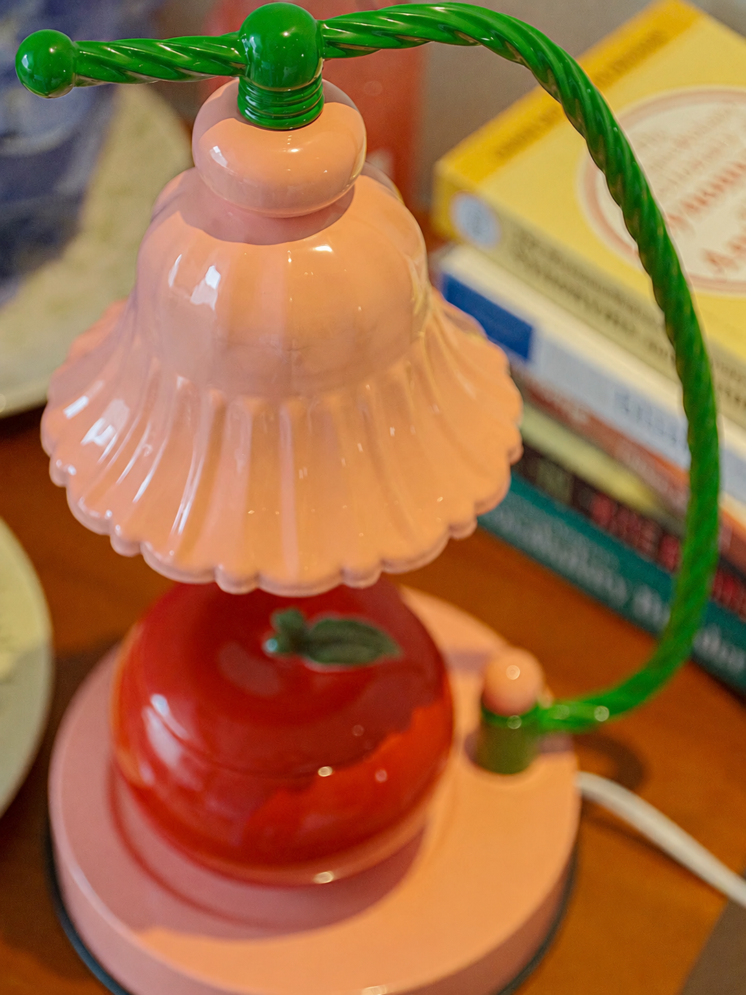 A retro-style table lamp with a pink bell-shaped shade, a red apple-shaped glass bulb, and a green twisted cord handle, shown on a wooden surface with books in the background.