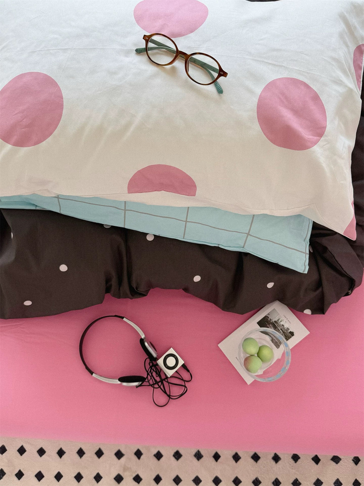 A white pillow with large pink dots rests over a blue checked pillow above the dark dotted cover and pink sheet.