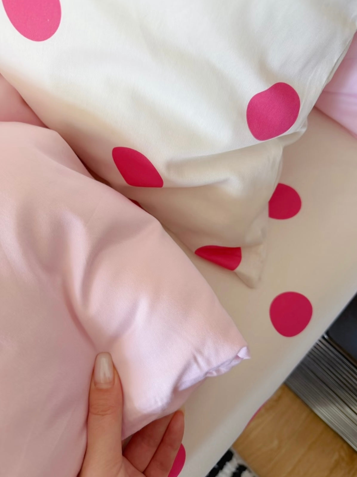 A hand holds plain pink bedding fabric beside a white pillowcase with large fuchsia dots.