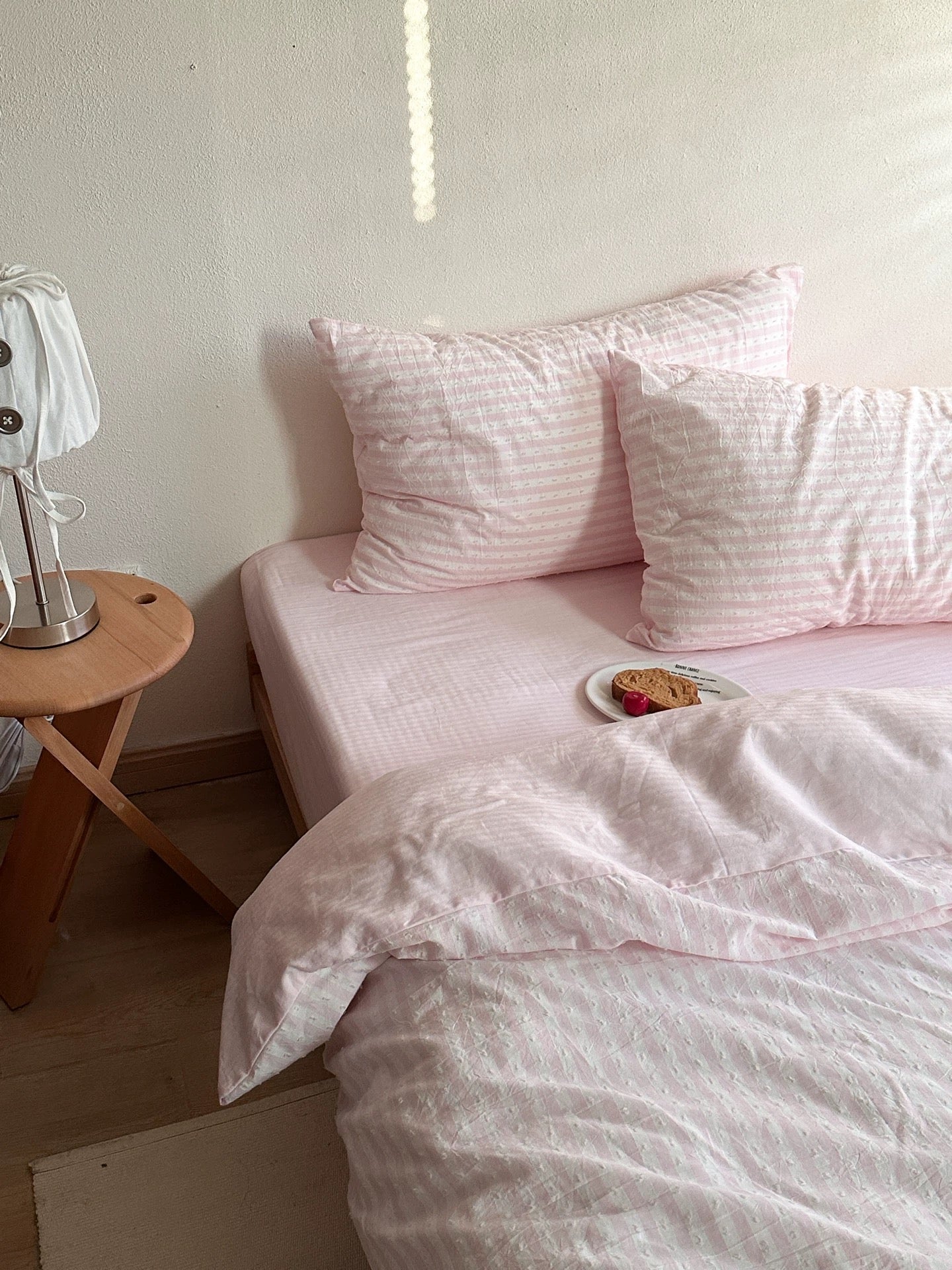 A neatly made bed with light pink bedding and matching pillows, styled in a bedroom setting with a small wooden side table and a lamp on the left.