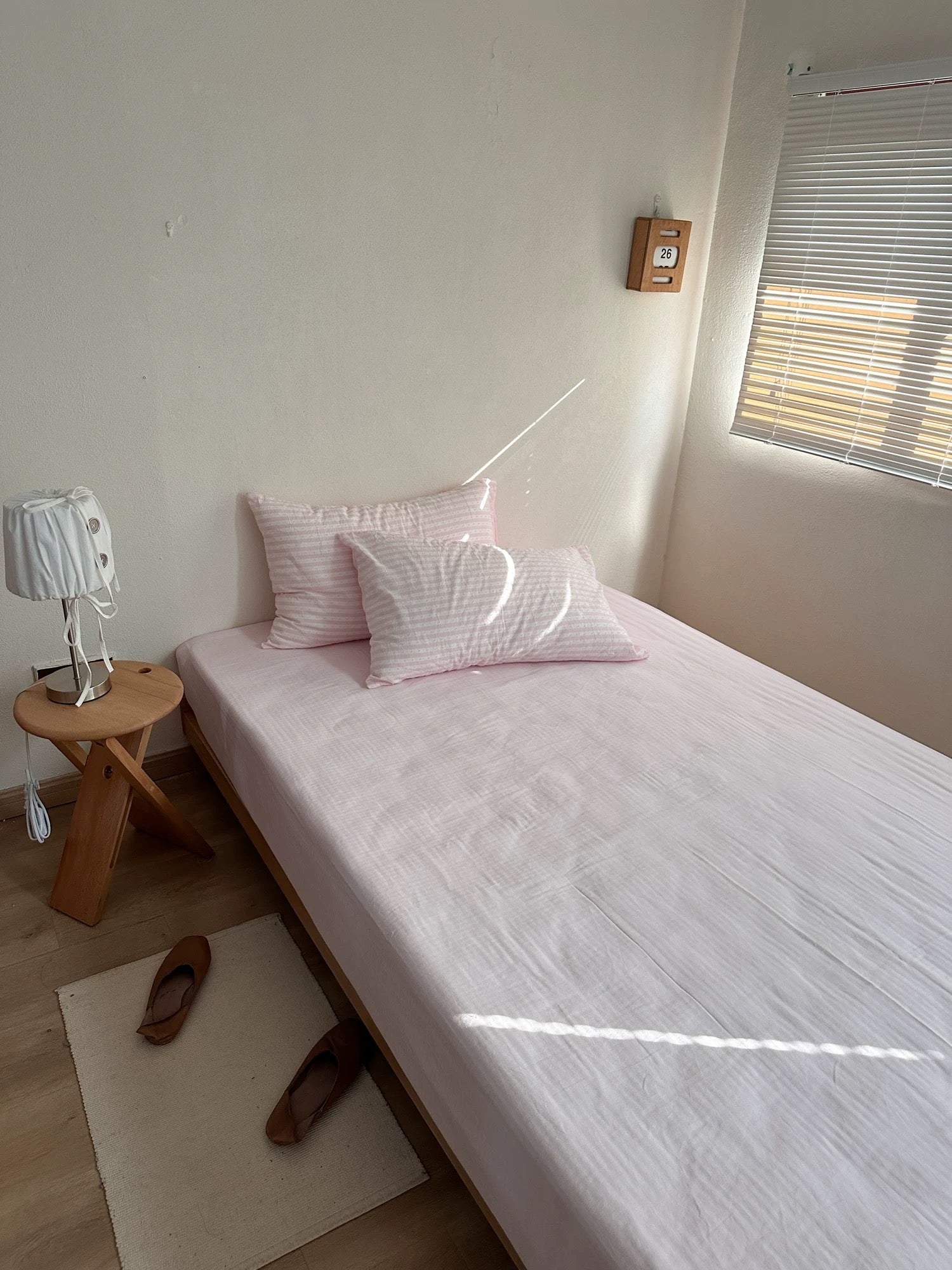 A single bed dressed in light pink striped bedding and matching pillows, placed in a minimalist bedroom with a small wooden side table, lamp, window blinds, and a pair of brown shoes on a rug.