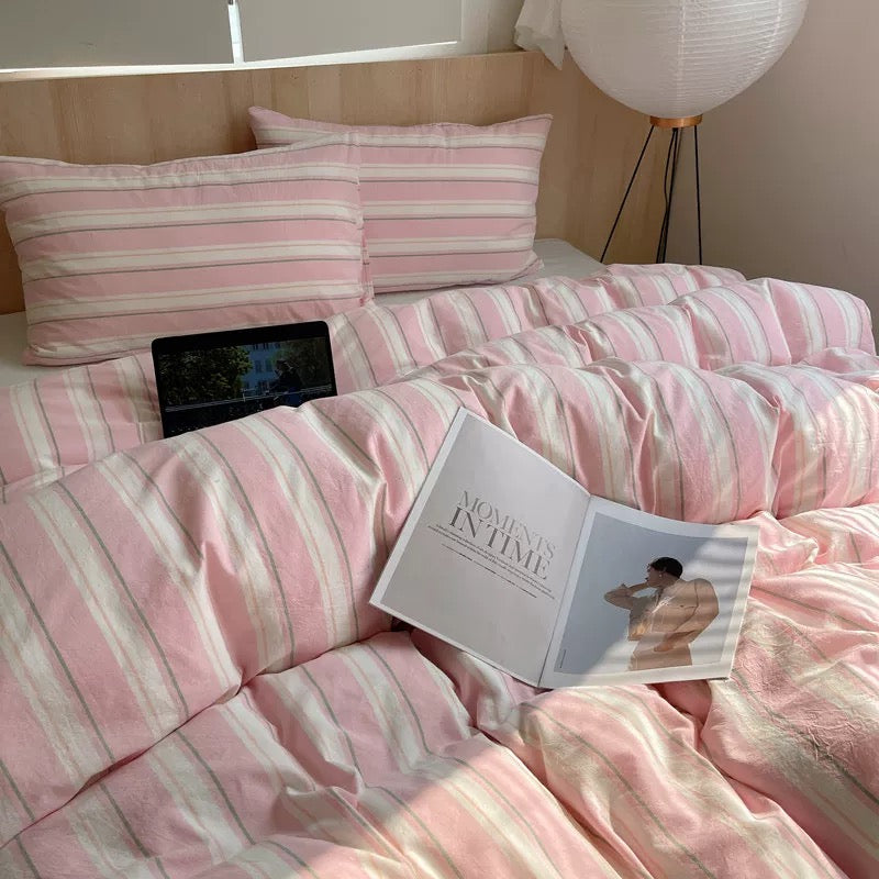 Pink and white striped bedding set on a bed with a nightstand and lamp in the background.
