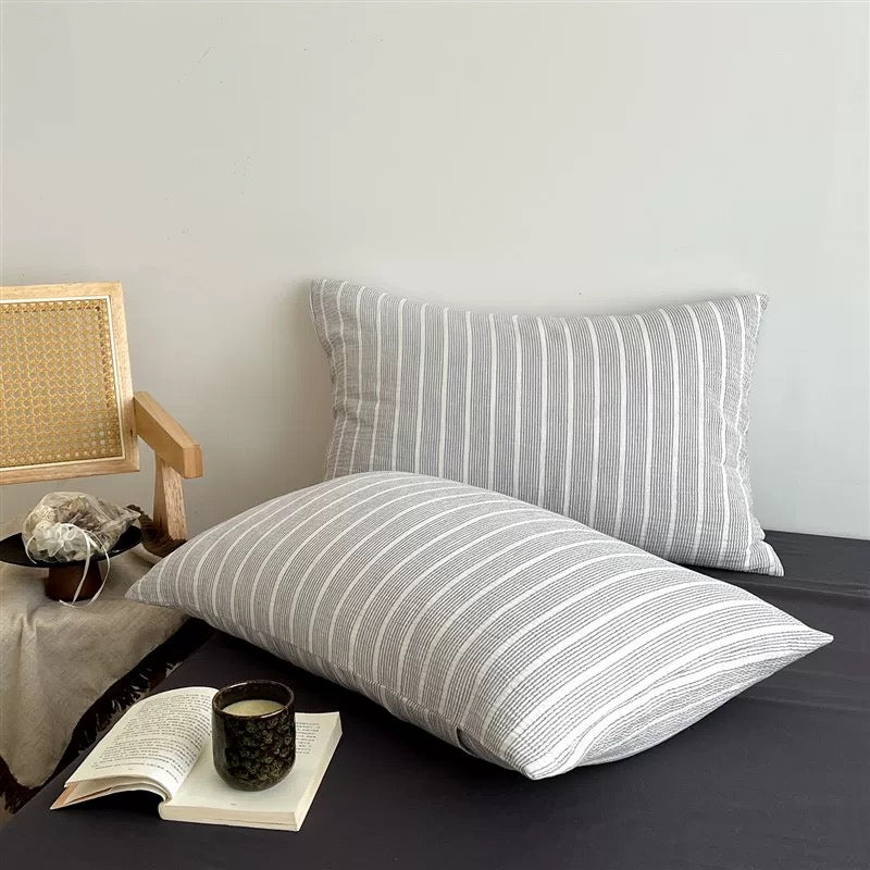 Two rectangular throw pillows with a light gray and white vertical stripe pattern, styled on a dark bed with a book, a cup, and a cane-back chair against a plain light gray wall.
