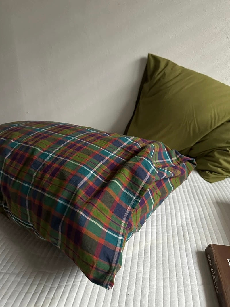 A multicolor plaid pillowcase in front of an olive green pillowcase on a white bed.
