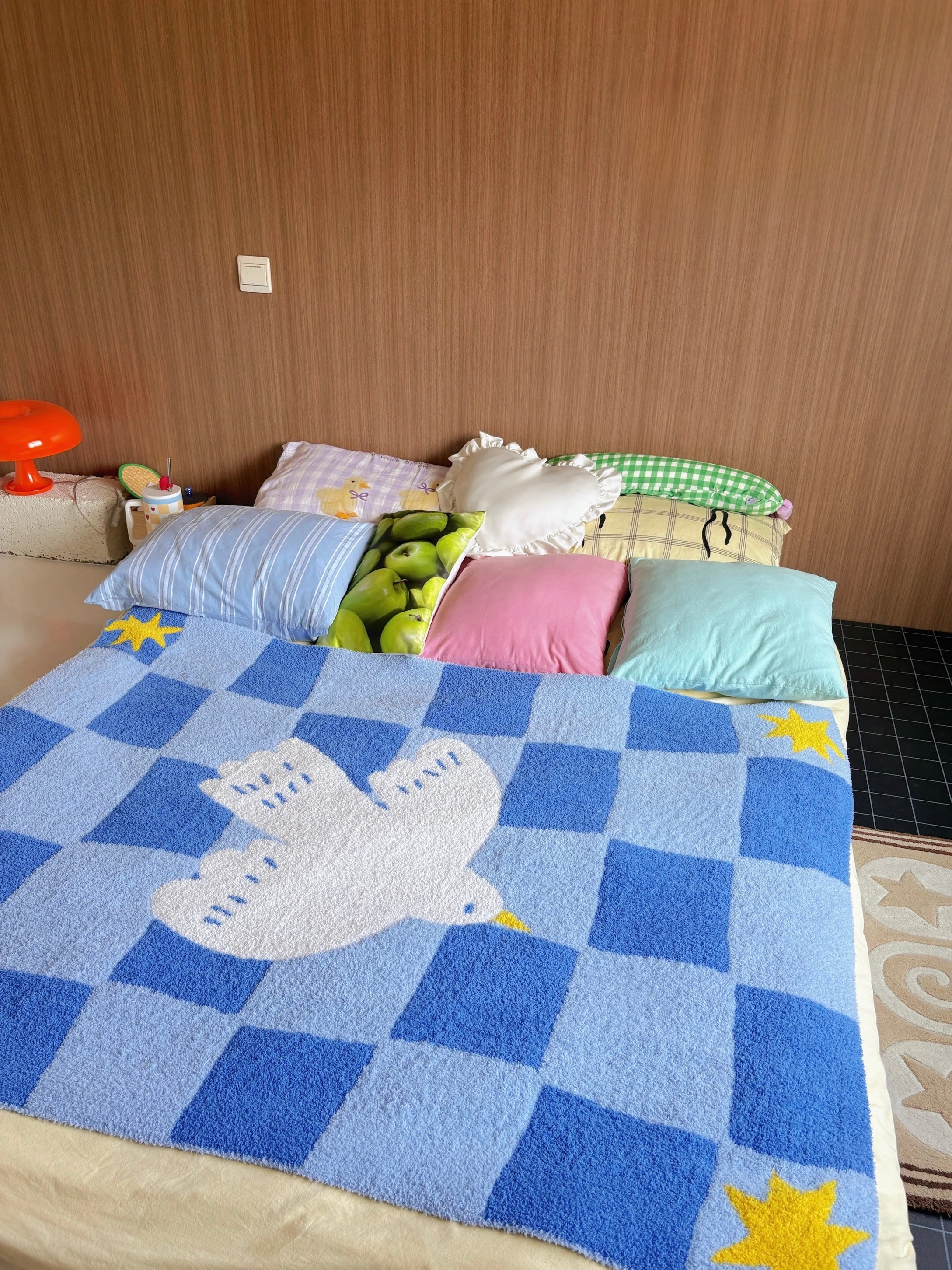 A blue and light blue checkered fleece blanket with a white dove design and yellow star accents, spread across a bed with colorful pillows in a bedroom setting.