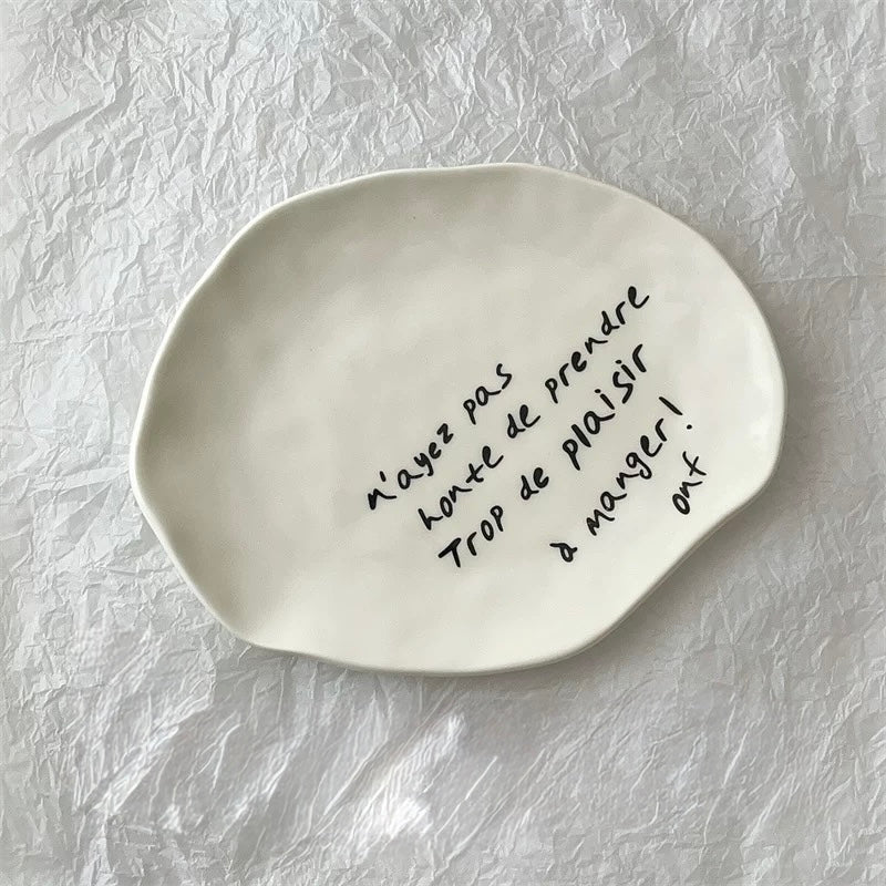 An organic-shaped off-white ceramic plate with black handwritten French text, displayed on a crumpled white paper background.