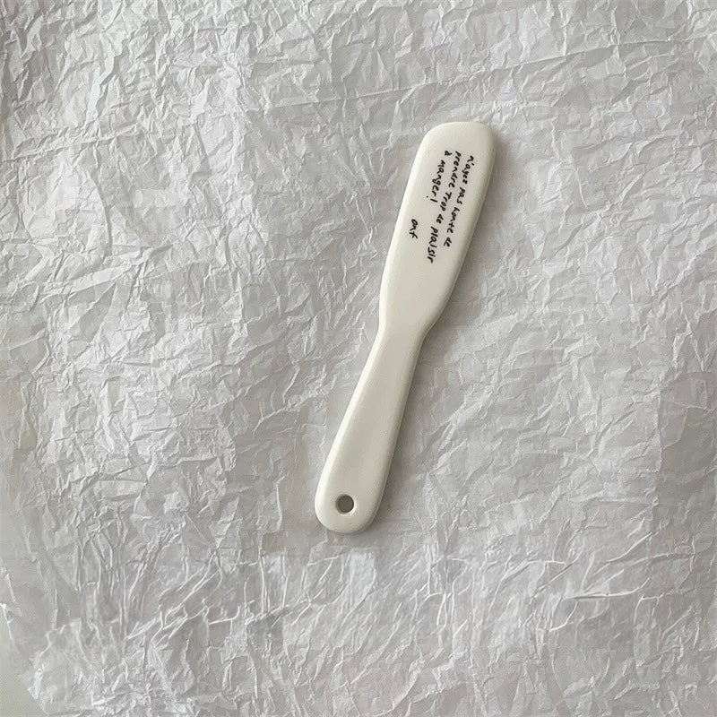 A white ceramic hair comb with black printed text on its head, resting on a textured, crumpled white paper background.