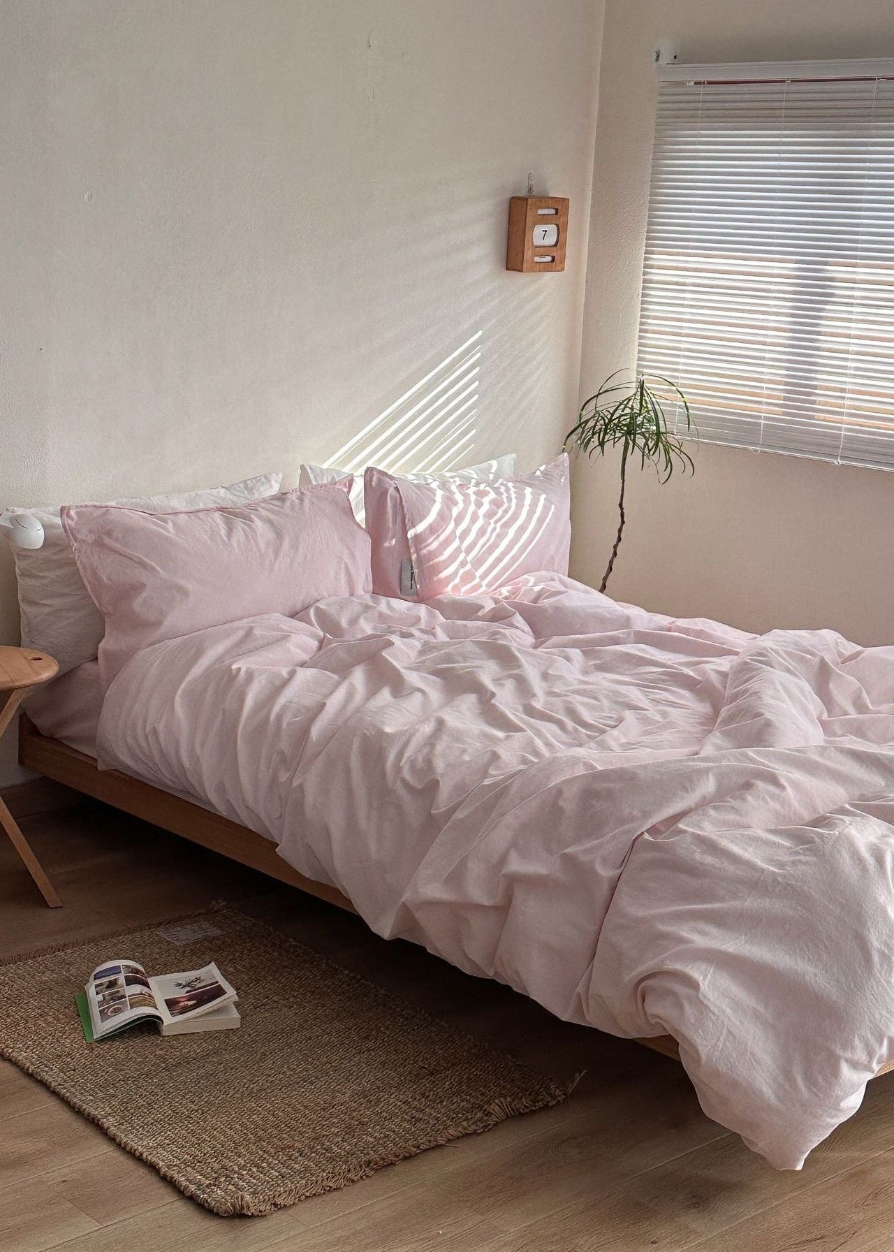 A bed dressed in a soft blush pink bedding set, styled in a minimalist bedroom with a wooden floor, a small side table, and a window with blinds.