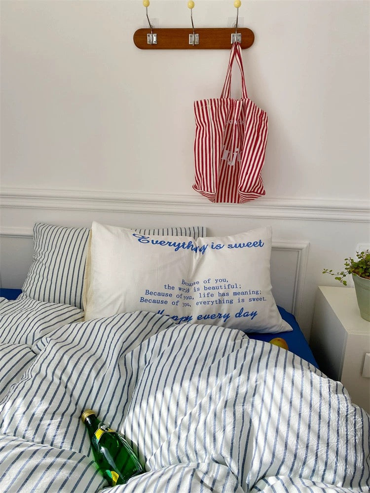 Bed with striped bedding and a pillow with text, red and white bag hanging above, white wall background