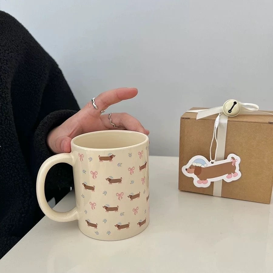 A cream-colored ceramic mug with a dachshund and bow pattern, held by a hand wearing a black sweater. A brown kraft gift box with a ribbon and sticker is on the table next to it.
